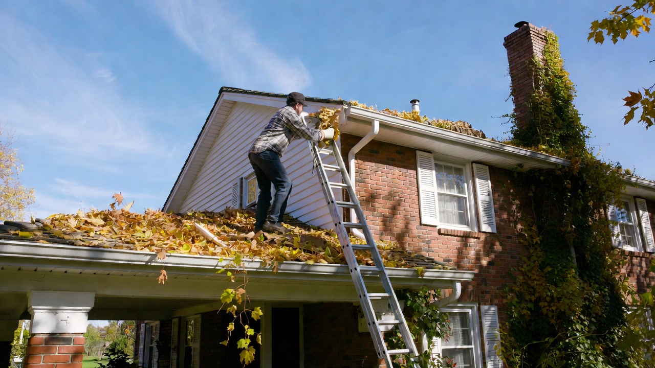 A cozy suburban home in early fall with a homeowner wearing gloves and using a ladder to clean gutters, surrounded by colorful autumn
