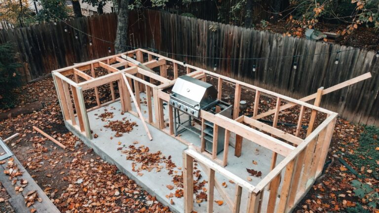 how to build an outdoor kitchen — An overhead view of a rustic backyard with a partially constructed outdoor kitchen showing exposed wooden framing, concrete foundation, and a stainless steel grill being installed, with autumn leaves scattered on the ground and string lights hung overhead, capturing the DIY work-in-progress phase