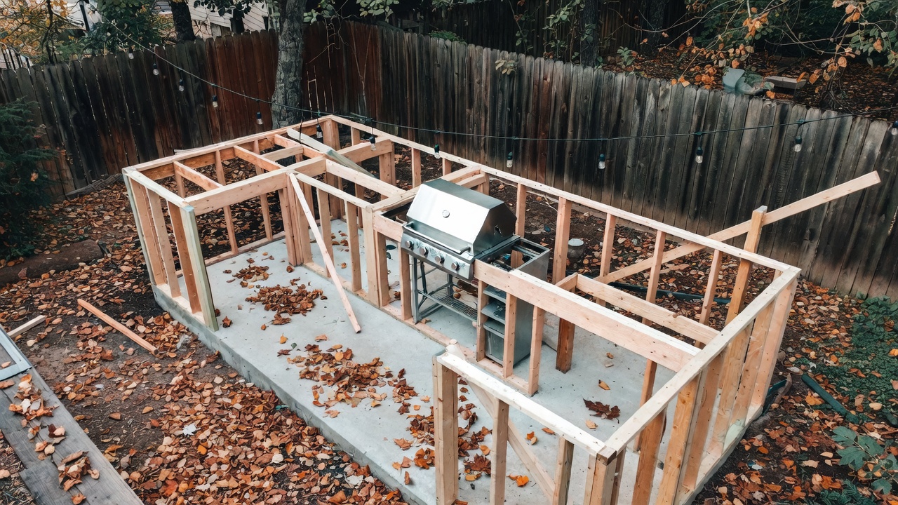 how to build an outdoor kitchen — An overhead view of a rustic backyard with a partially constructed outdoor kitchen showing exposed wooden framing, concrete foundation, and a stainless steel grill being installed, with autumn leaves scattered on the ground and string lights hung overhead, capturing the DIY work-in-progress phase